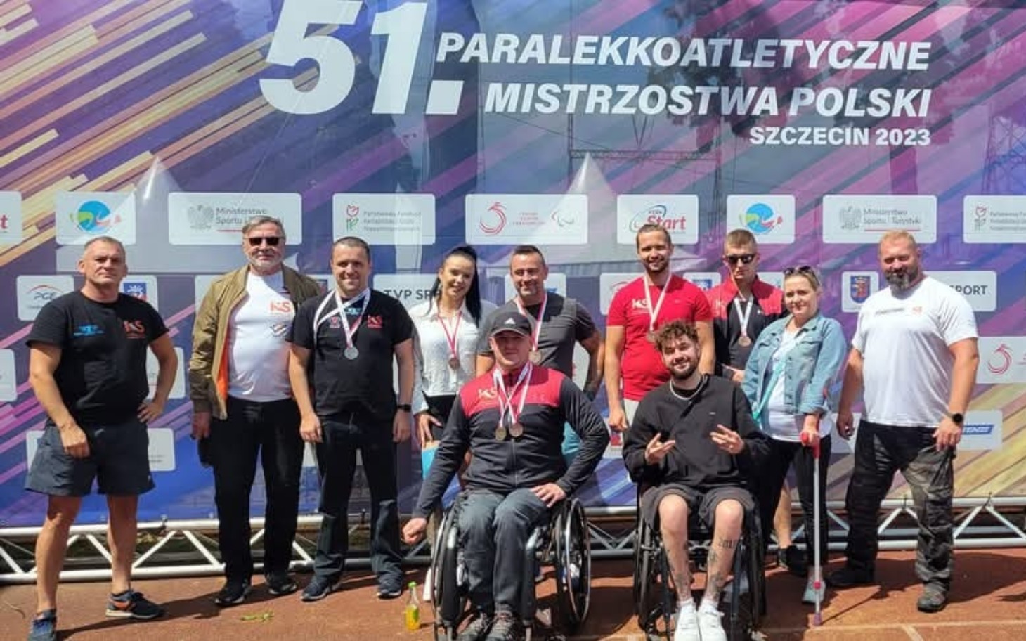 
        Grupa ludzi na zawodach paraolimpijskich, wszyscy uśmiechnięci.
    