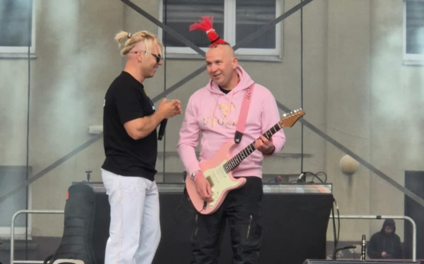 
        Dwóch mężczyzn na scenie, jeden z gitarą, rozmawiających.
    