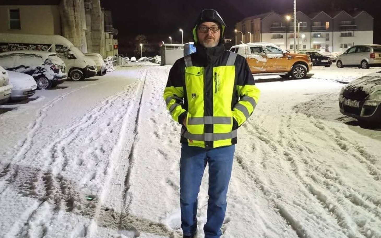 
        Mężczyzna w odblaskowej kurtce na zaśnieżonym parkingu.
    