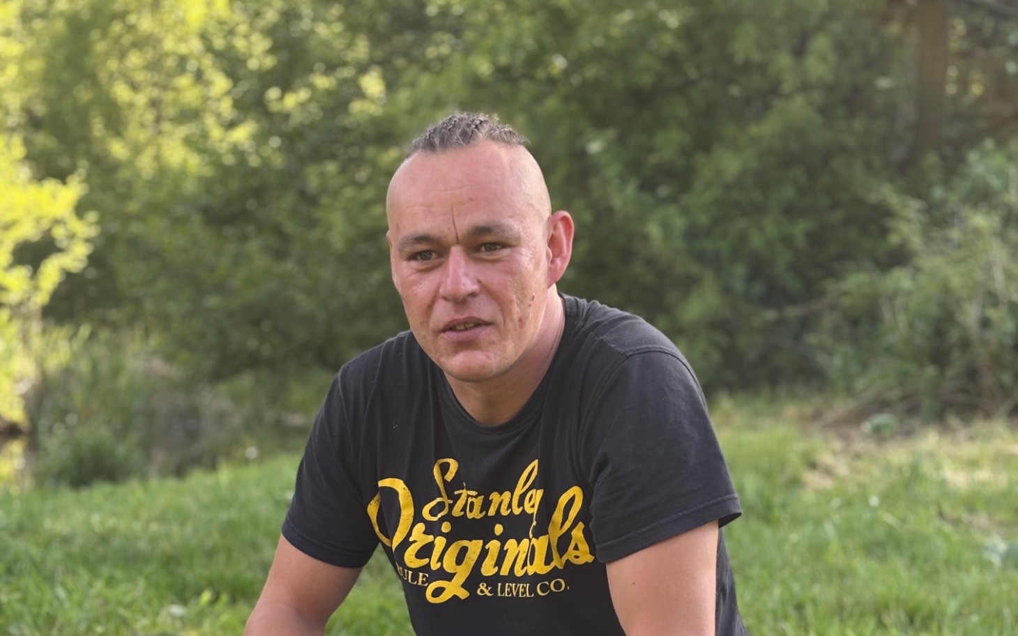 
        Mężczyzna siedzący na trawie, w tle zieleń.
    