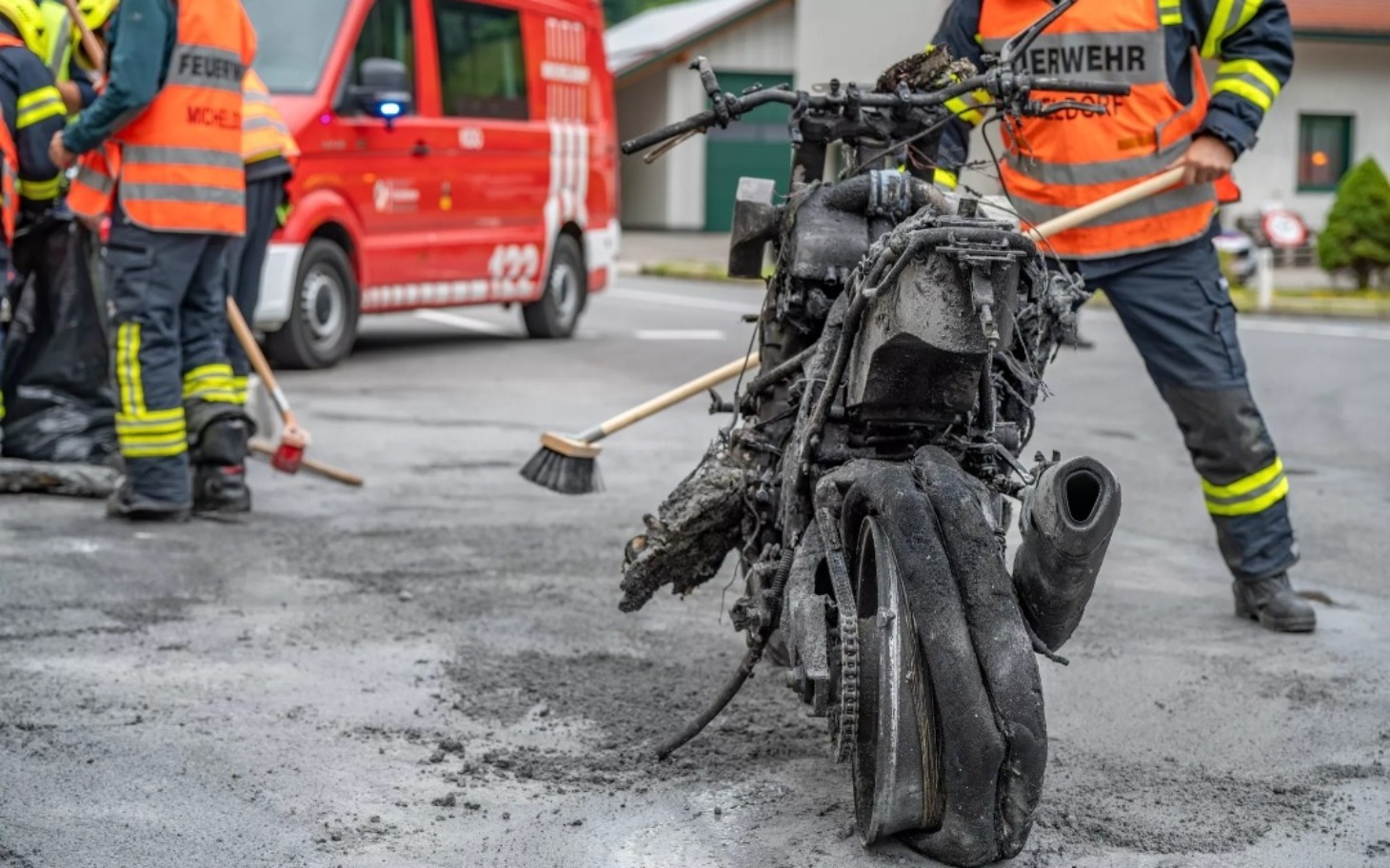 
        Zniszczony motocykl po pożarze, obok strażacy w akcji.
    