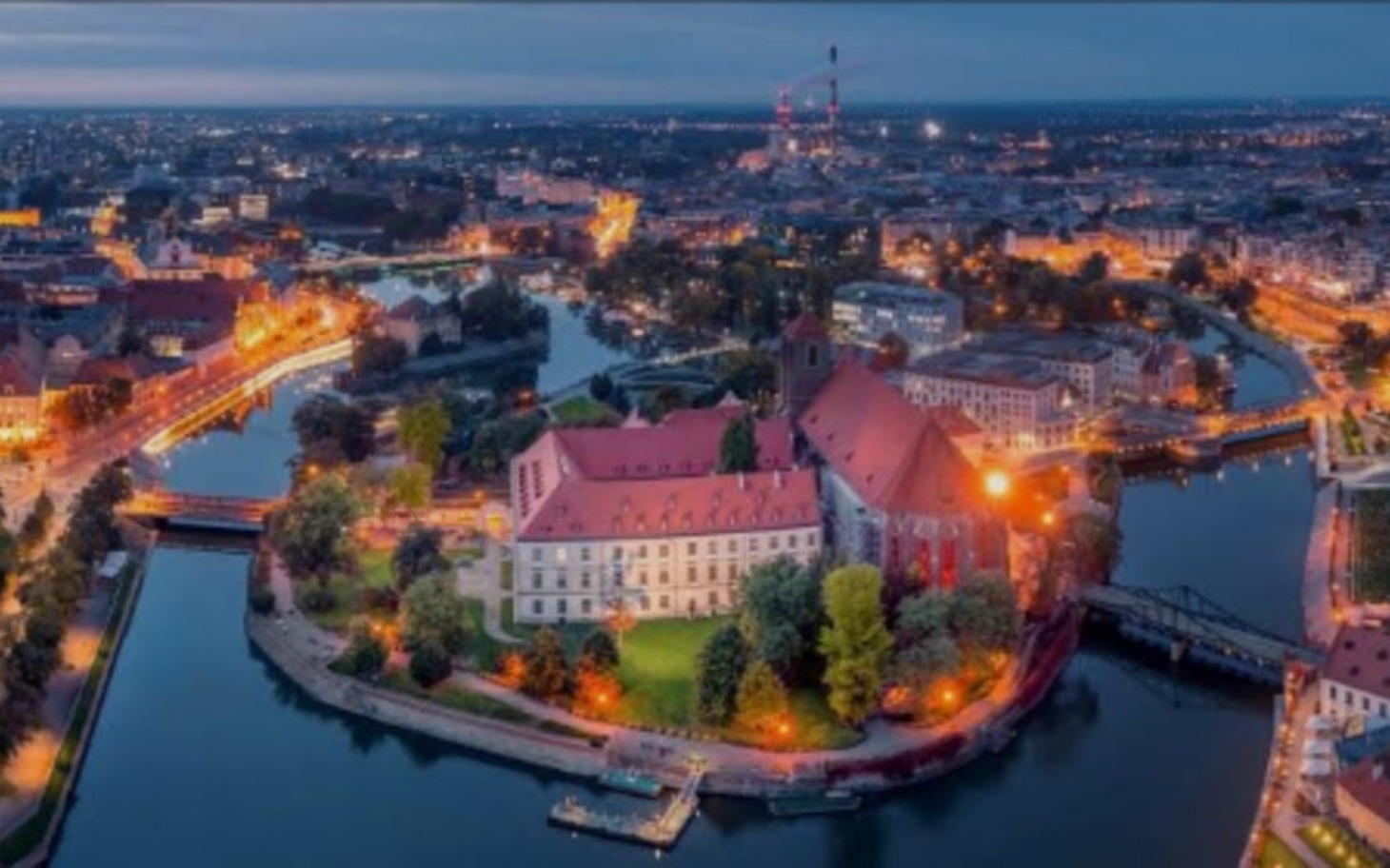 
        Widok na miasto nocą, z oświetlonymi budynkami i rzeką.
    