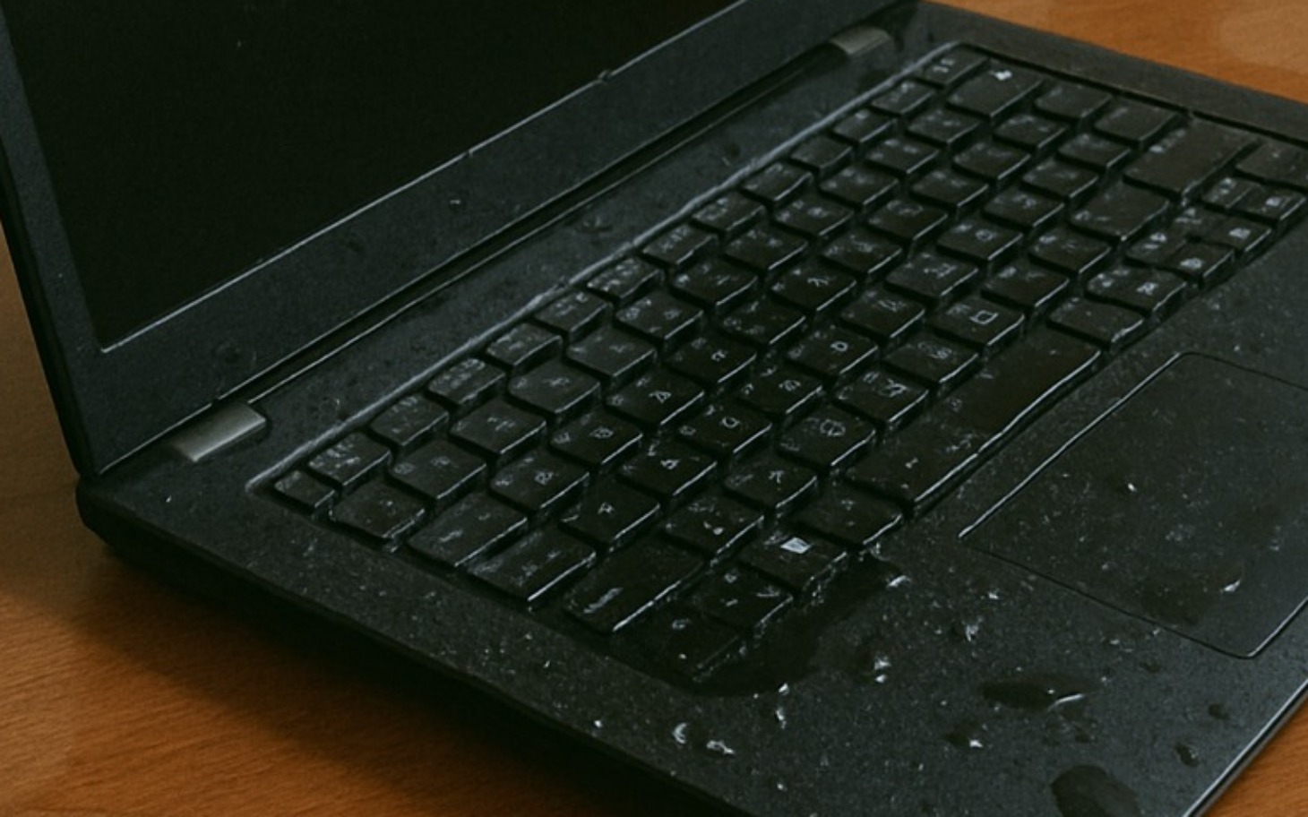 
        Zalany laptop na drewnianym biurku, widoczne krople wody.
    