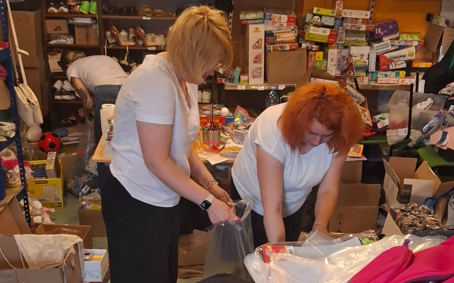 
        Dwie kobiety sortują rzeczy w pomieszczeniu pełnym darów.
    
