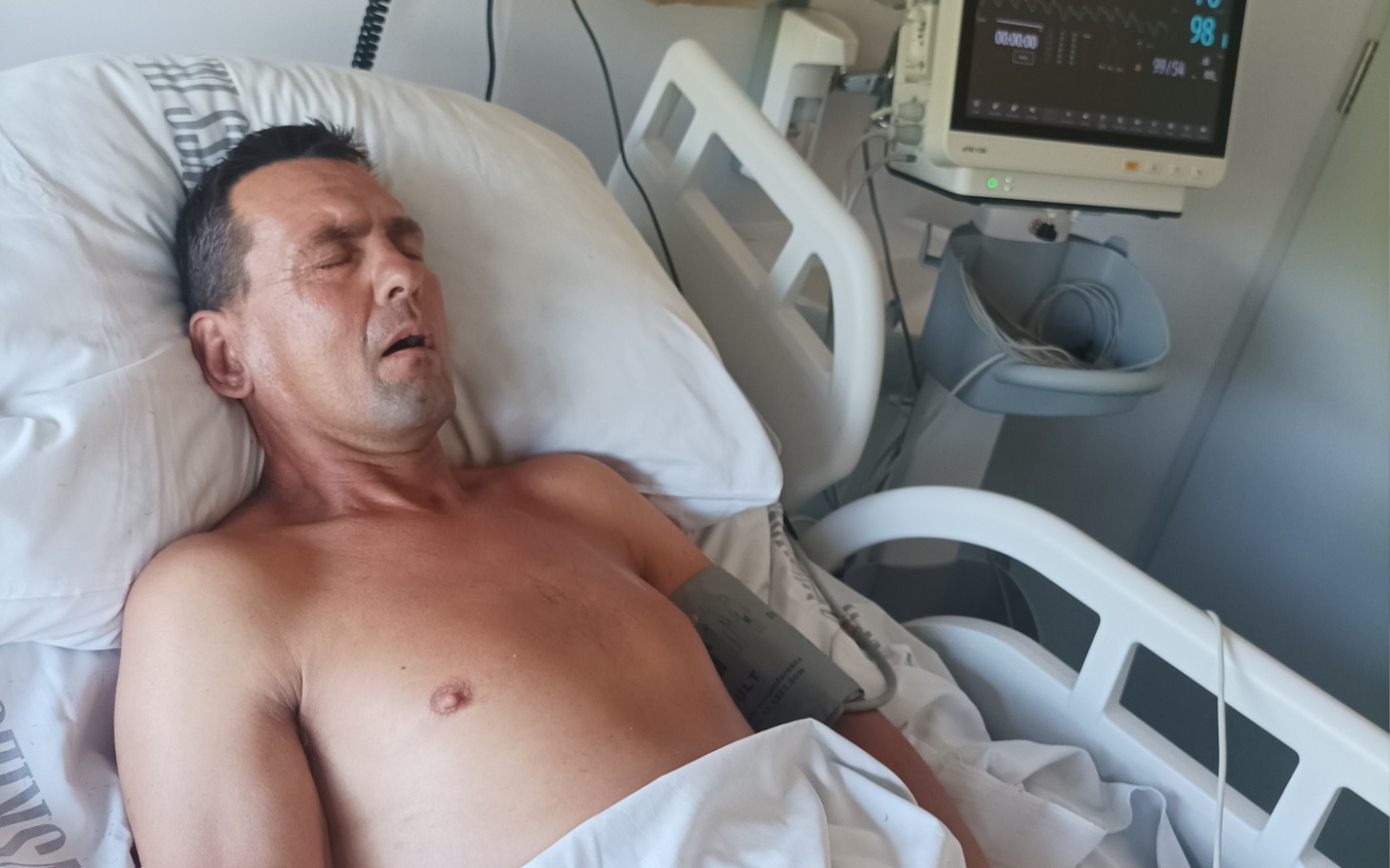 
        Mężczyzna leżący w szpitalnym łóżku, z monitorami wokół.
    