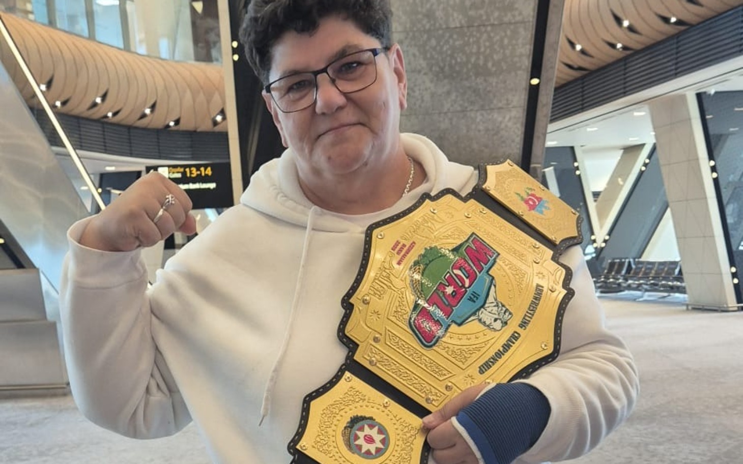 
        Kobieta z pasem mistrza, uśmiechnięta, w hali lotniska.
    