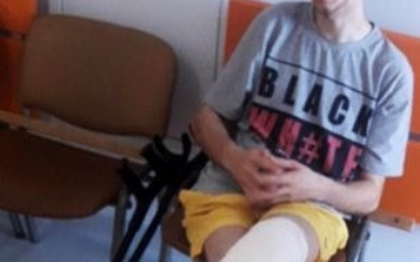 
        Młody mężczyzna w szarej koszulce siedzi na krześle, z opatrunkiem na nodze.
    