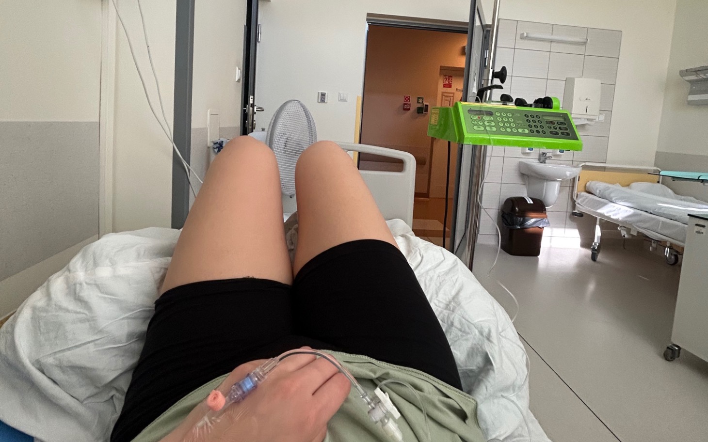 
        Osoba w szpitalu, leżąca na łóżku, z widokiem na salę i sprzęt medyczny.
    