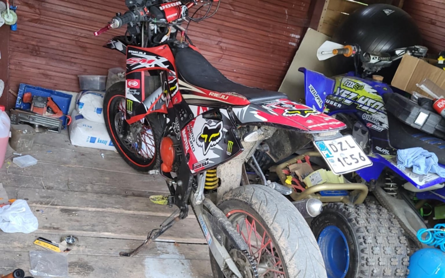 
        Motocykl w garażu, z widocznymi naklejkami i częściami.
    