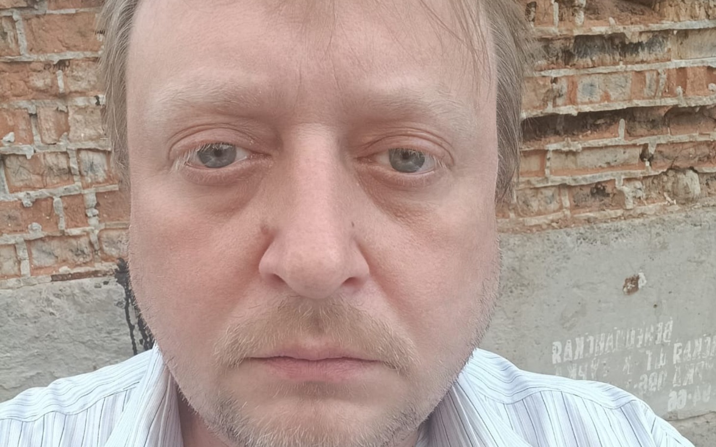 
        Mężczyzna z poważnym wyrazem twarzy, siedzący na tle ceglanej ściany.
    
