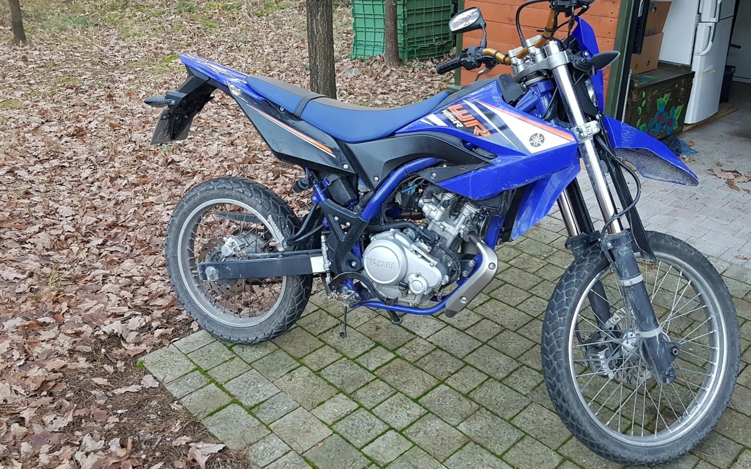 
        Niebieski motocykl terenowy na tle liści.
    