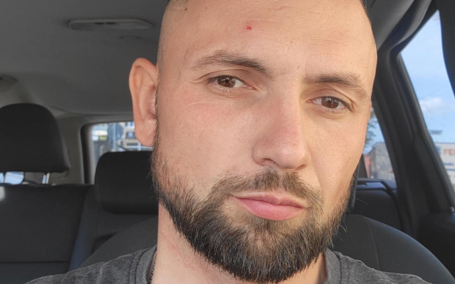 
        Mężczyzna z brodą w samochodzie, patrzący w kamerę.
    