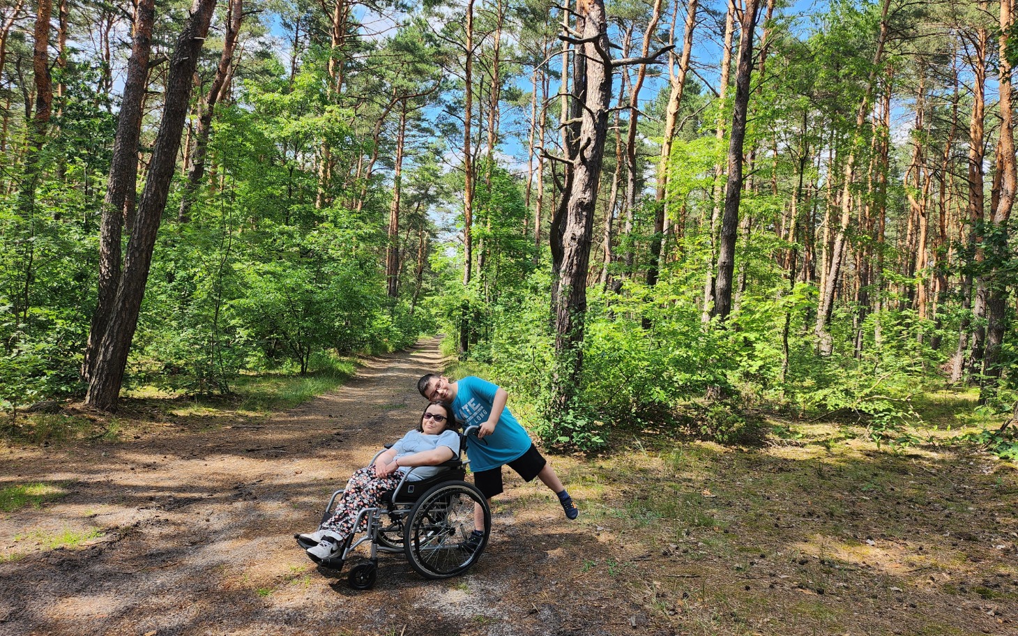 
        Osoba na wózku inwalidzkim w lesie, z uśmiechniętym towarzyszem.
    