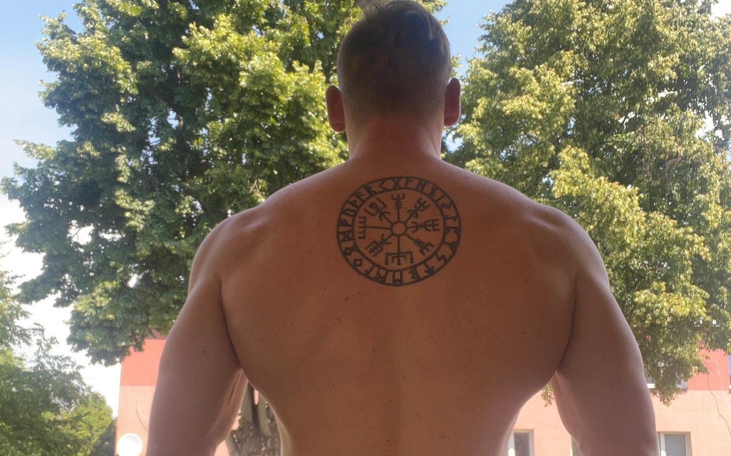 
        Mężczyzna z tatuażem na plecach, stojący na tle drzew.
    