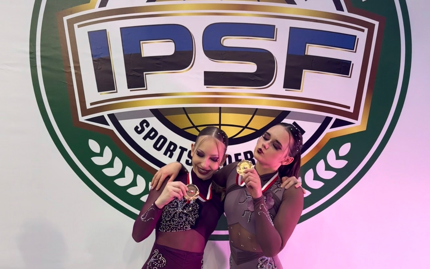
        Dwie dziewczyny z medalami, pozujące na tle logo IPSF.
    