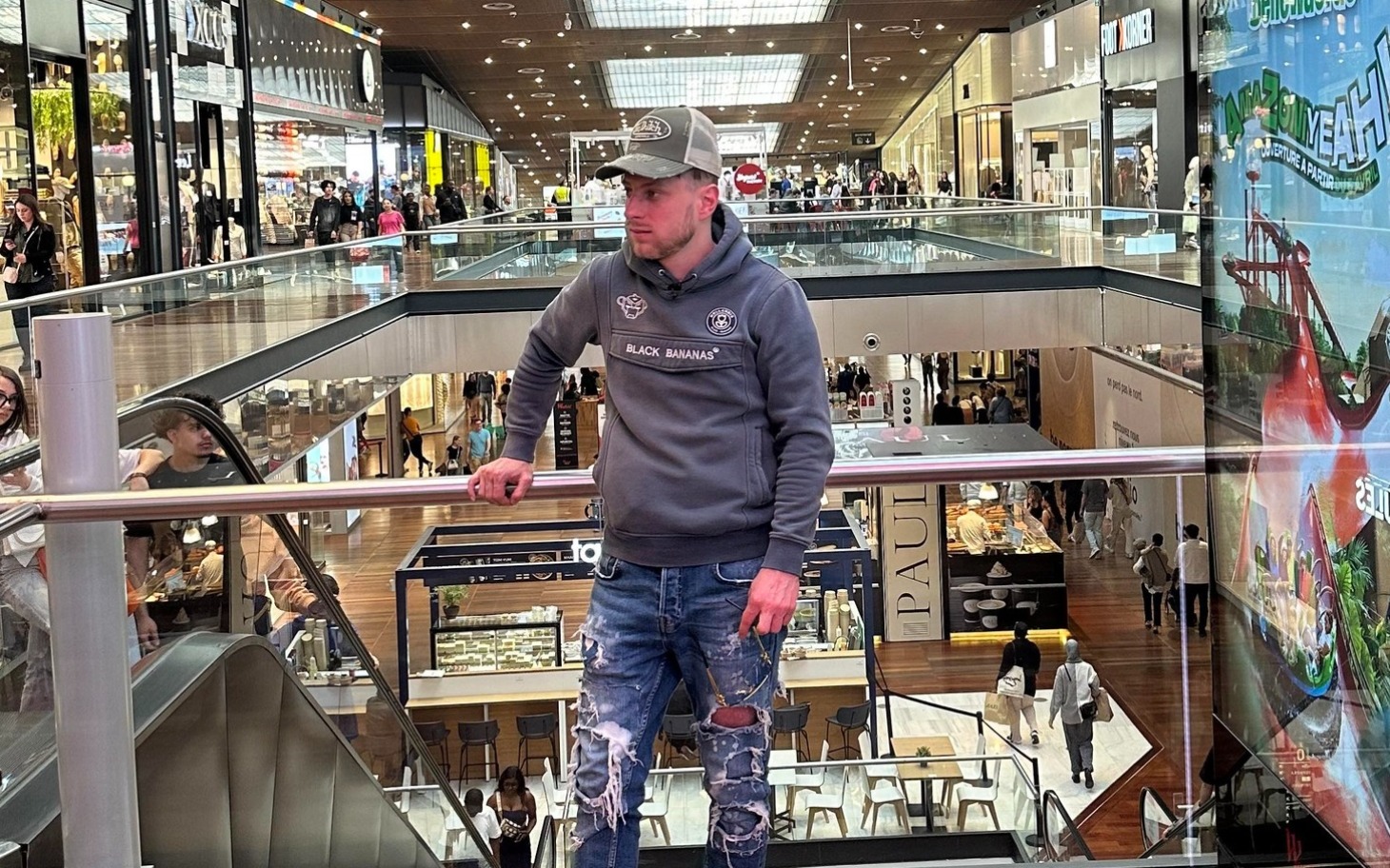 
        Mężczyzna w szarym swetrze w centrum handlowym, otoczony ludźmi.
    