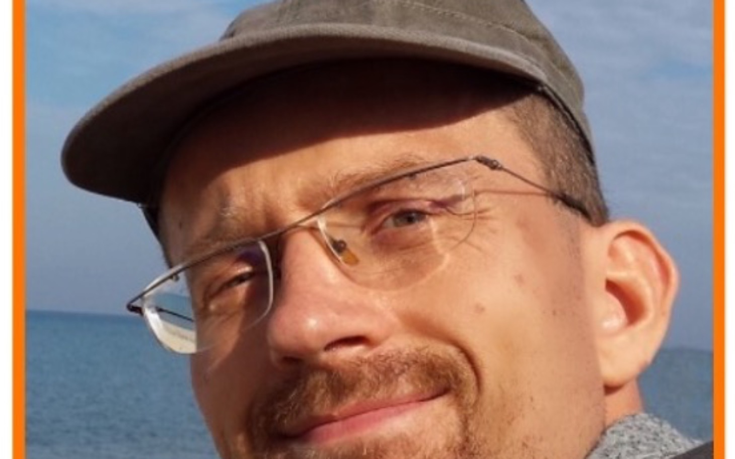 
        Mężczyzna w okularach, uśmiechający się na tle morza.
    