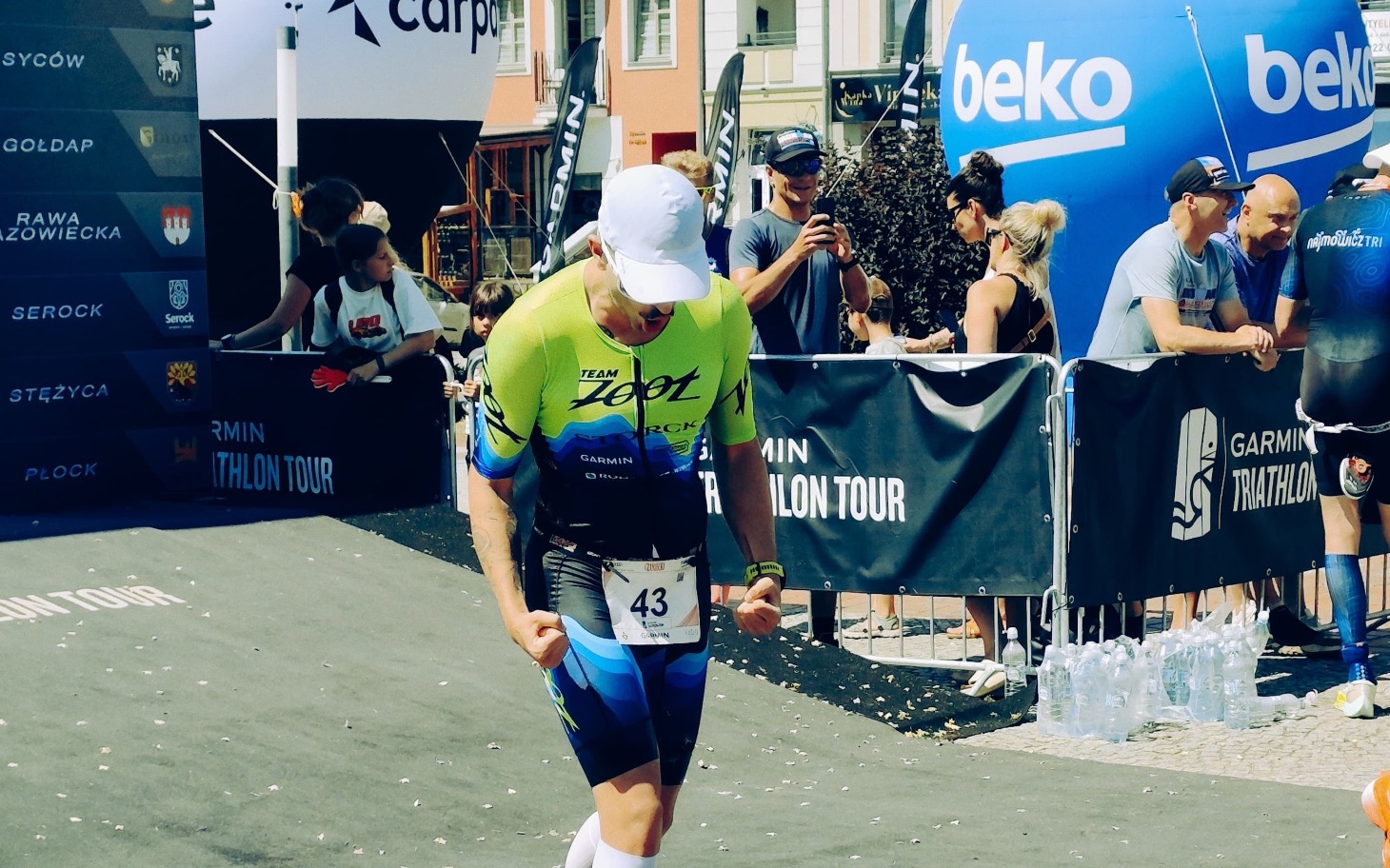 
        Zawodnik w stroju triathlonowym na mecie, w tle kibice.
    
