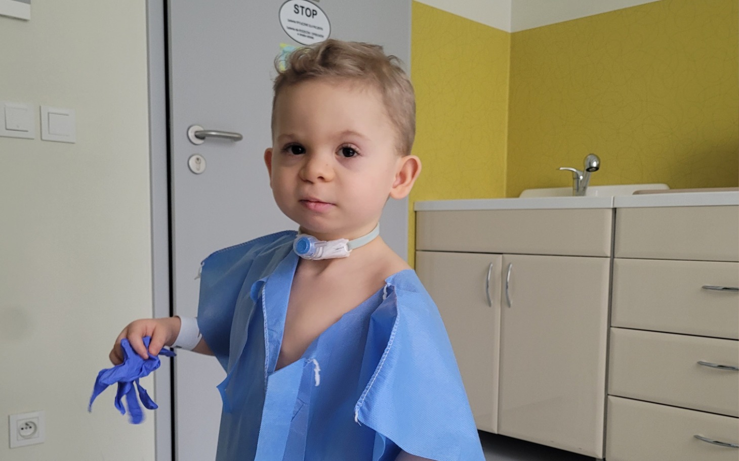 
        Mały chłopiec w szpitalnym fartuchu, trzymający rękawiczki.
    