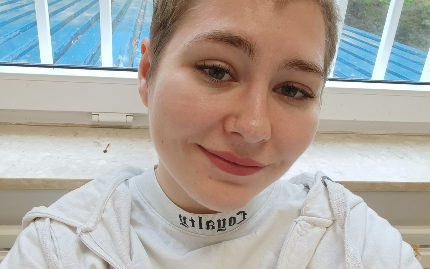 
        Młoda osoba uśmiechająca się do kamery w jasnym pomieszczeniu.
    