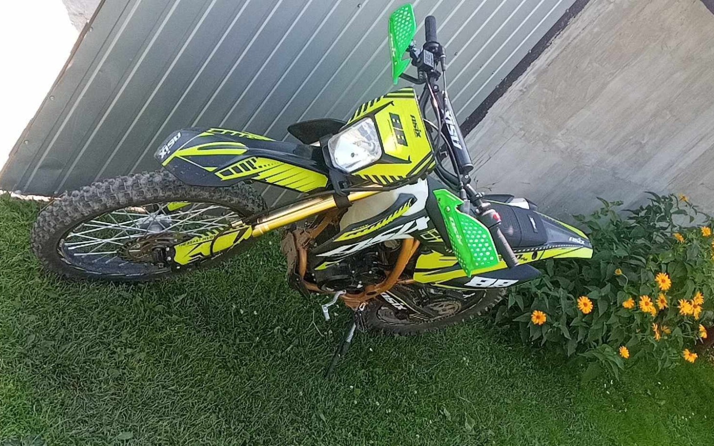 
        Motocykl terenowy w ogrodzie, otoczony kwiatami.
    