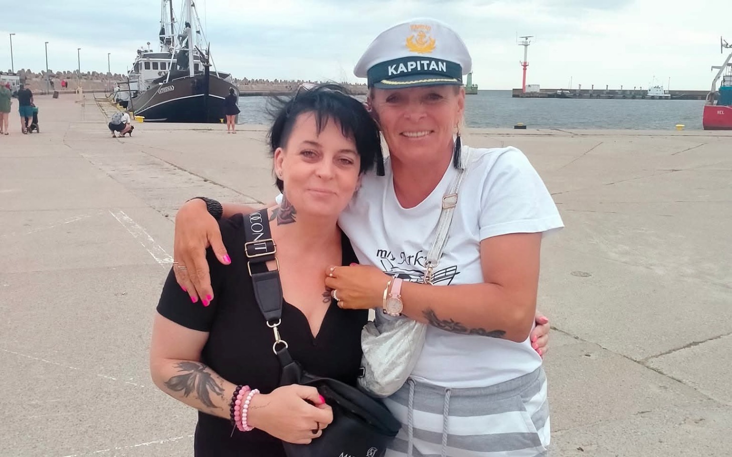 
        Dwie kobiety przytulają się na tle portu, jedna w kapitańskiej czapce.
    
