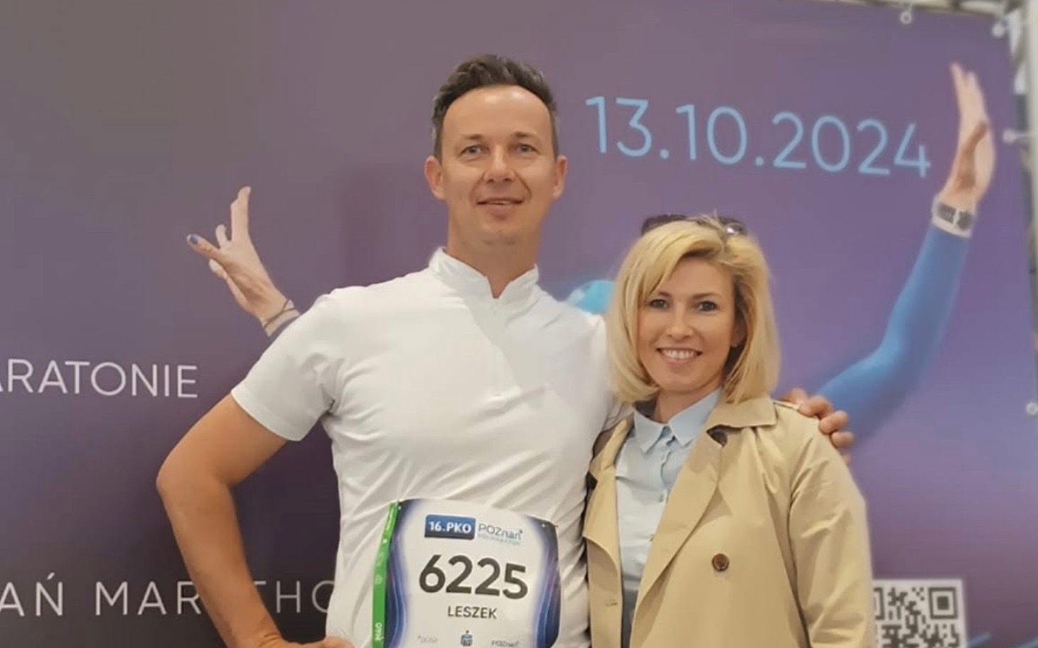 
        Para na tle plakatu maratonu, mężczyzna w koszulce startowej.
    