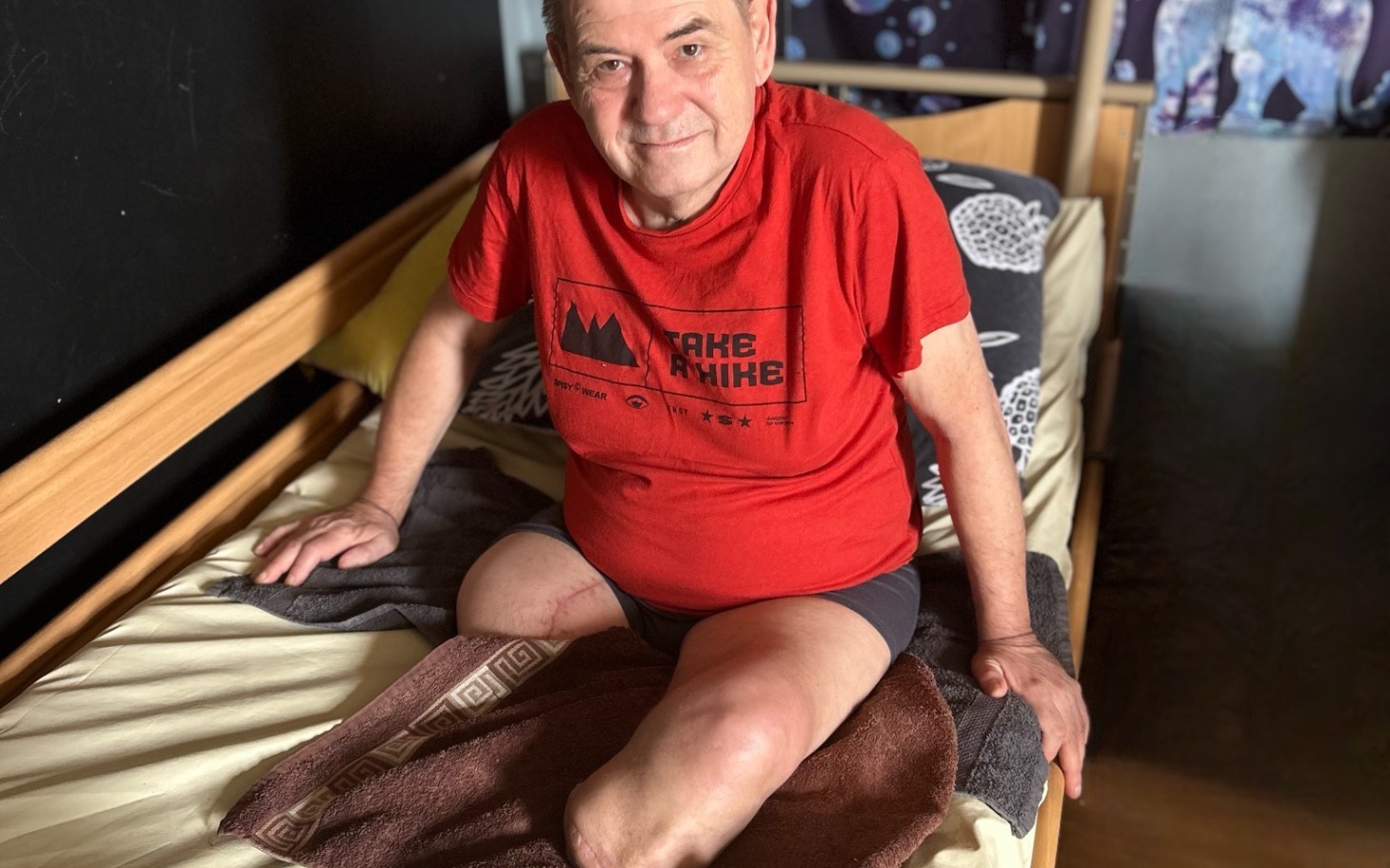 
        Starszy mężczyzna siedzący na łóżku, w czerwonym podkoszulku.
    