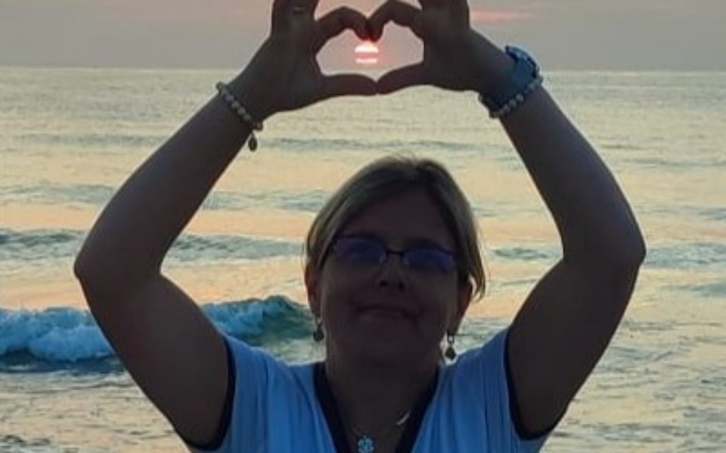 
        Kobieta na plaży tworzy serce z rąk, w tle zachód słońca.
    