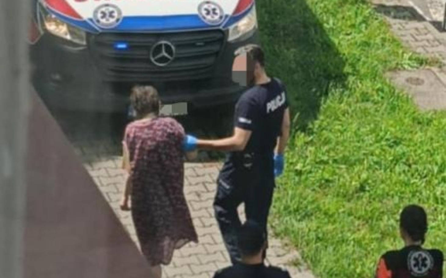 
        Policjant prowadzi starszą kobietę w sukience, obok zespół ratunkowy.
    
