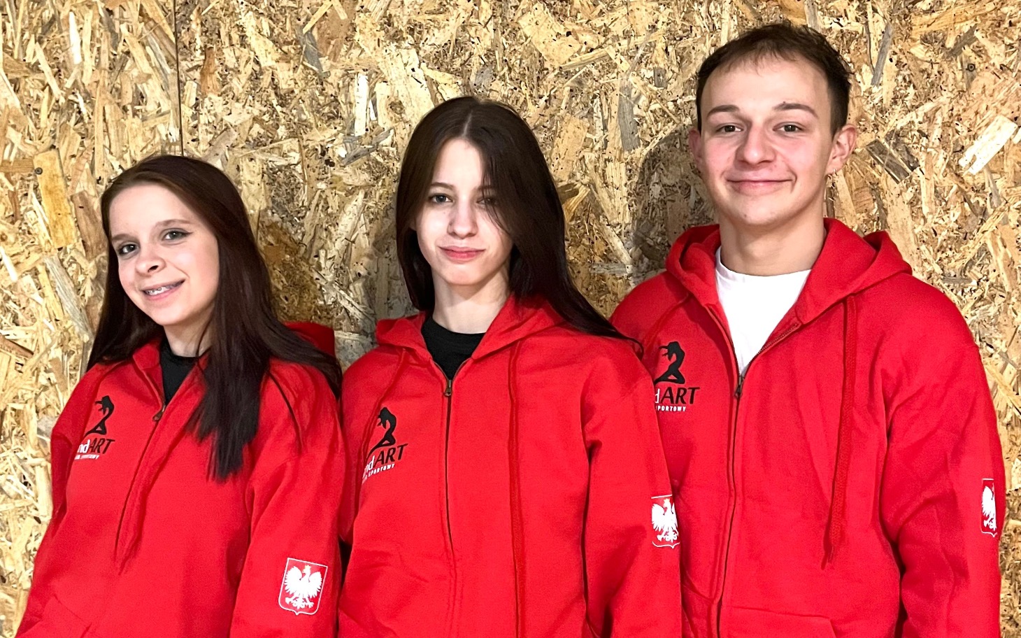 
        Trójka młodych ludzi w czerwonych bluzach na tle drewnianej ściany.
    