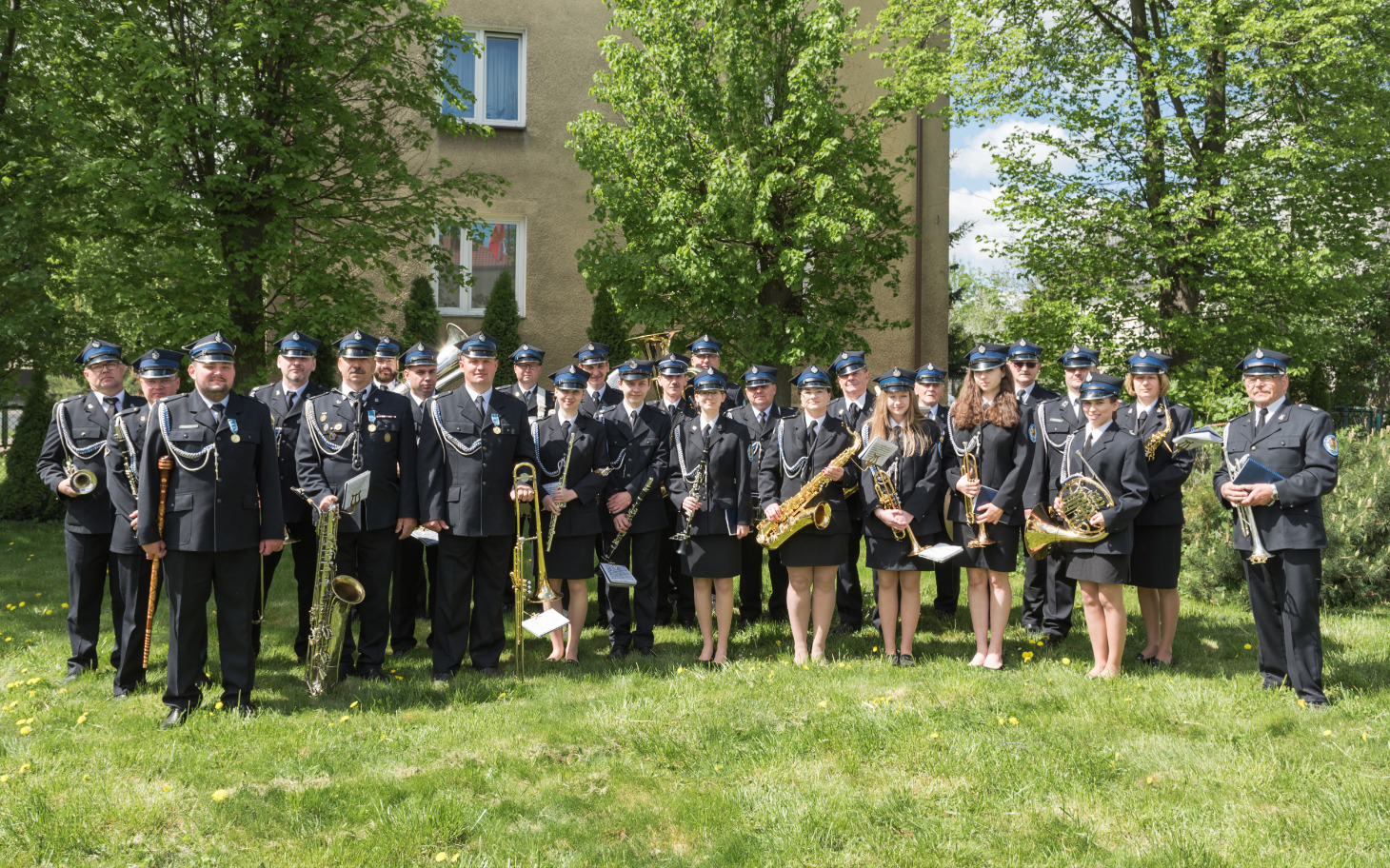 
        Orkiestra dęta w pełnym składzie, pozująca na tle zieleni.
    