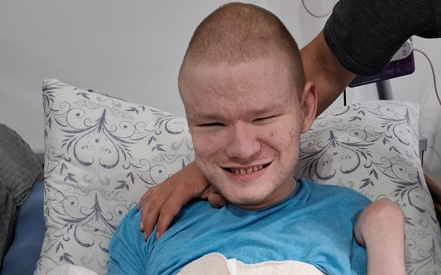 
        Młody mężczyzna uśmiechający się w szpitalnym łóżku, z opieką obok.
    