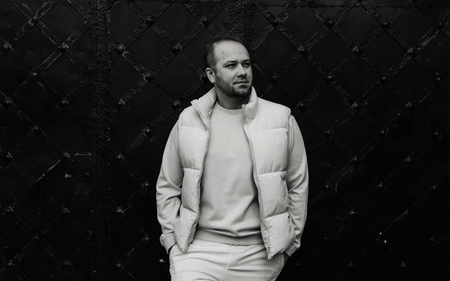 
        Mężczyzna w białej pikowanej kurtce na czarnym tle, minimalistyczny portret.
    