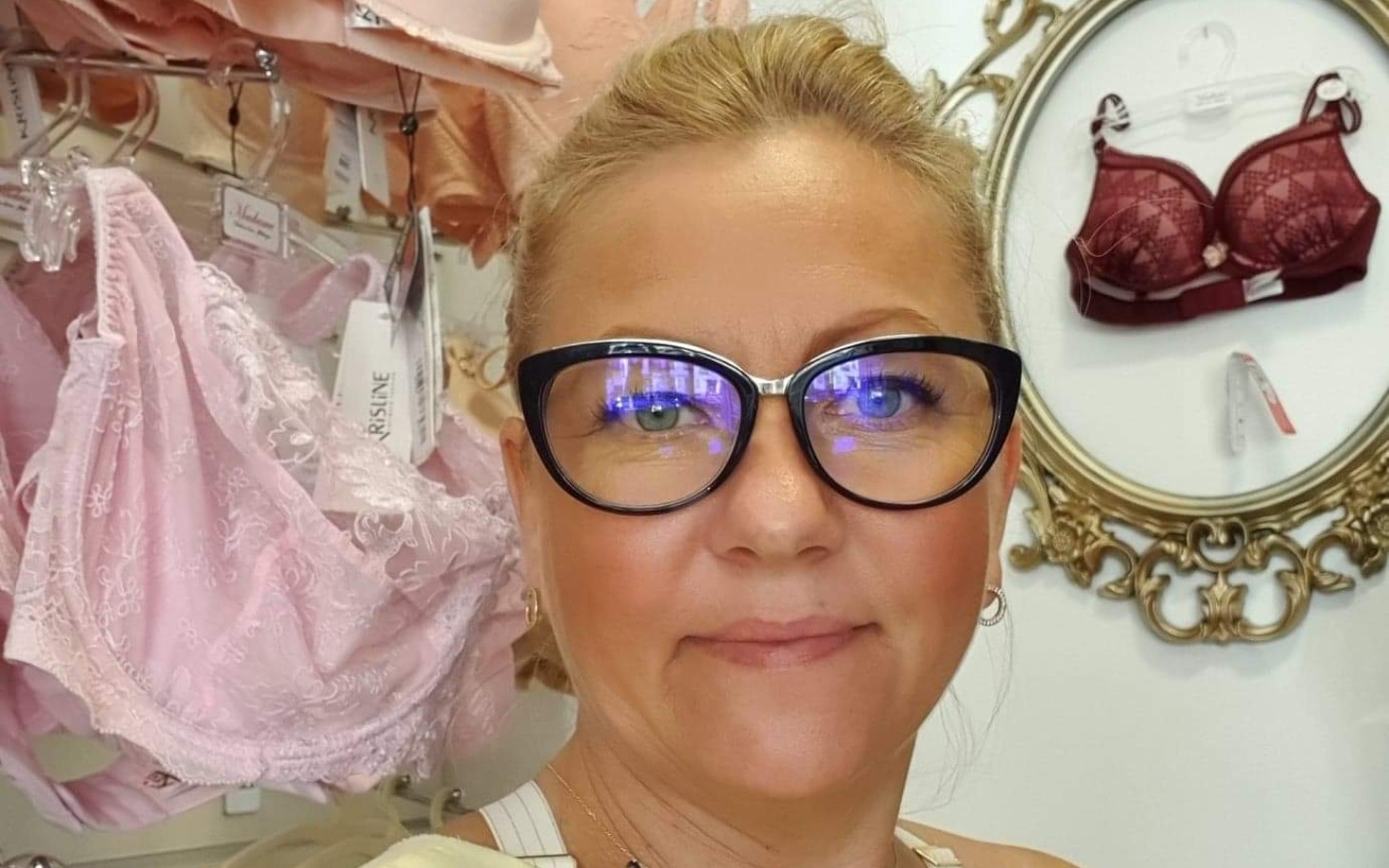 
        Kobieta w okularach stoi w sklepie z bielizną, uśmiechając się do kamery.
    
