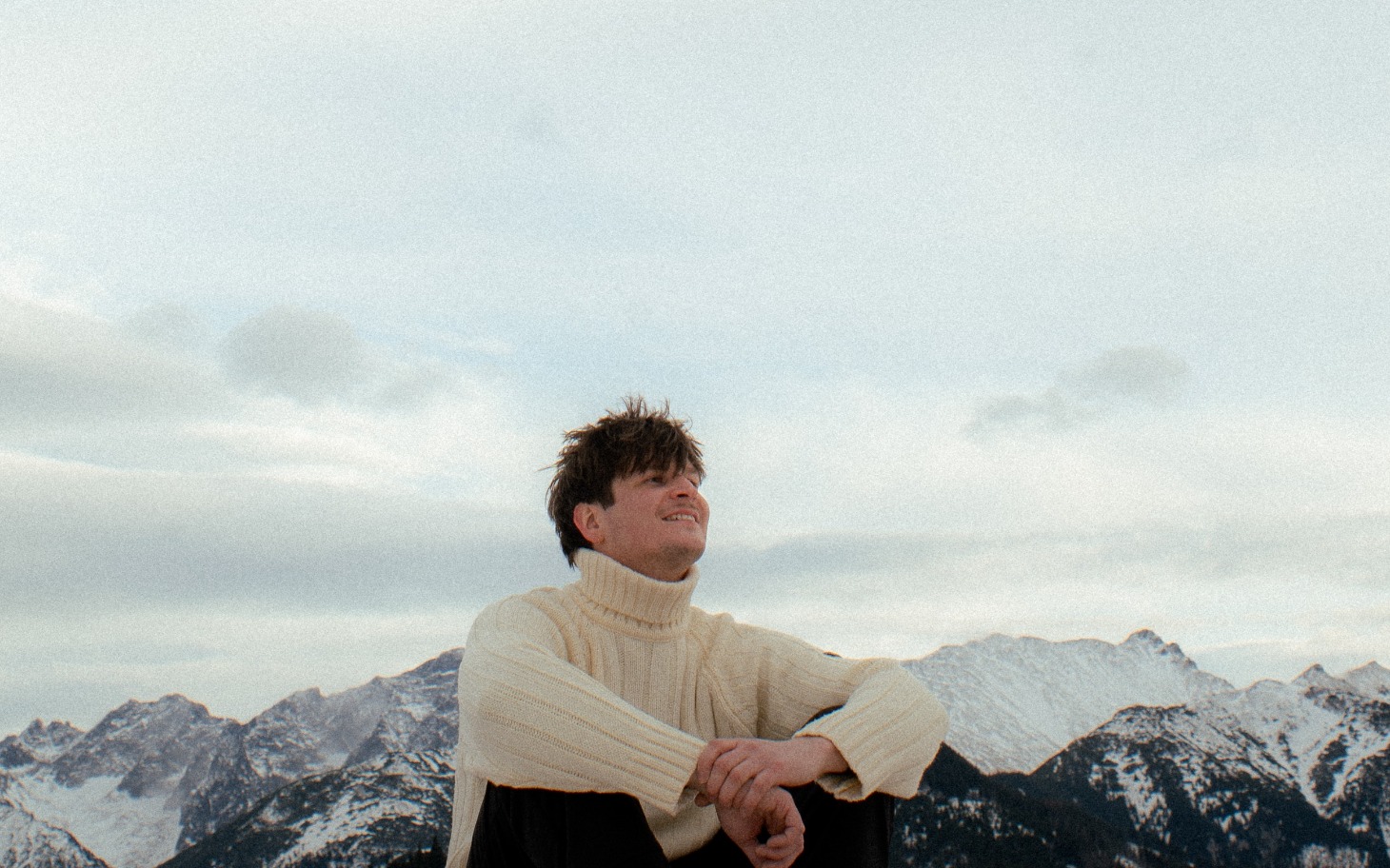 
        Mężczyzna w swetrze, siedzi na górze z widokiem na góry.
    