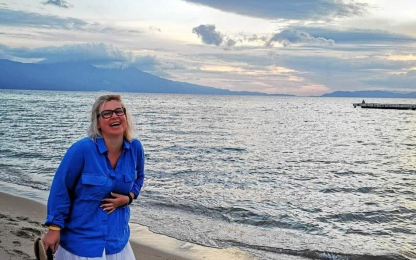 
        Kobieta uśmiechająca się na plaży, w tle morze i góry.
    
