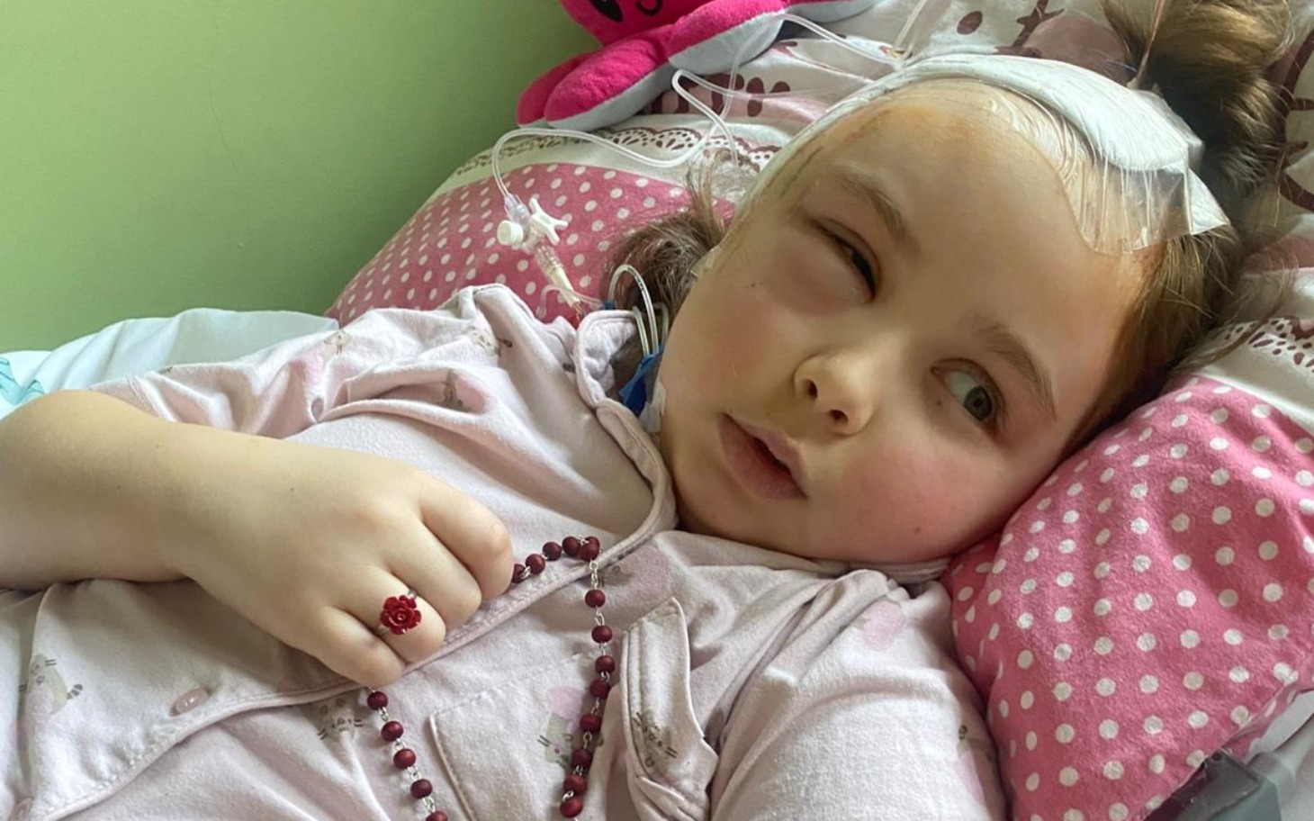 
        Mała dziewczynka leży w łóżku z opaską na głowie i różowymi poduszkami.
    