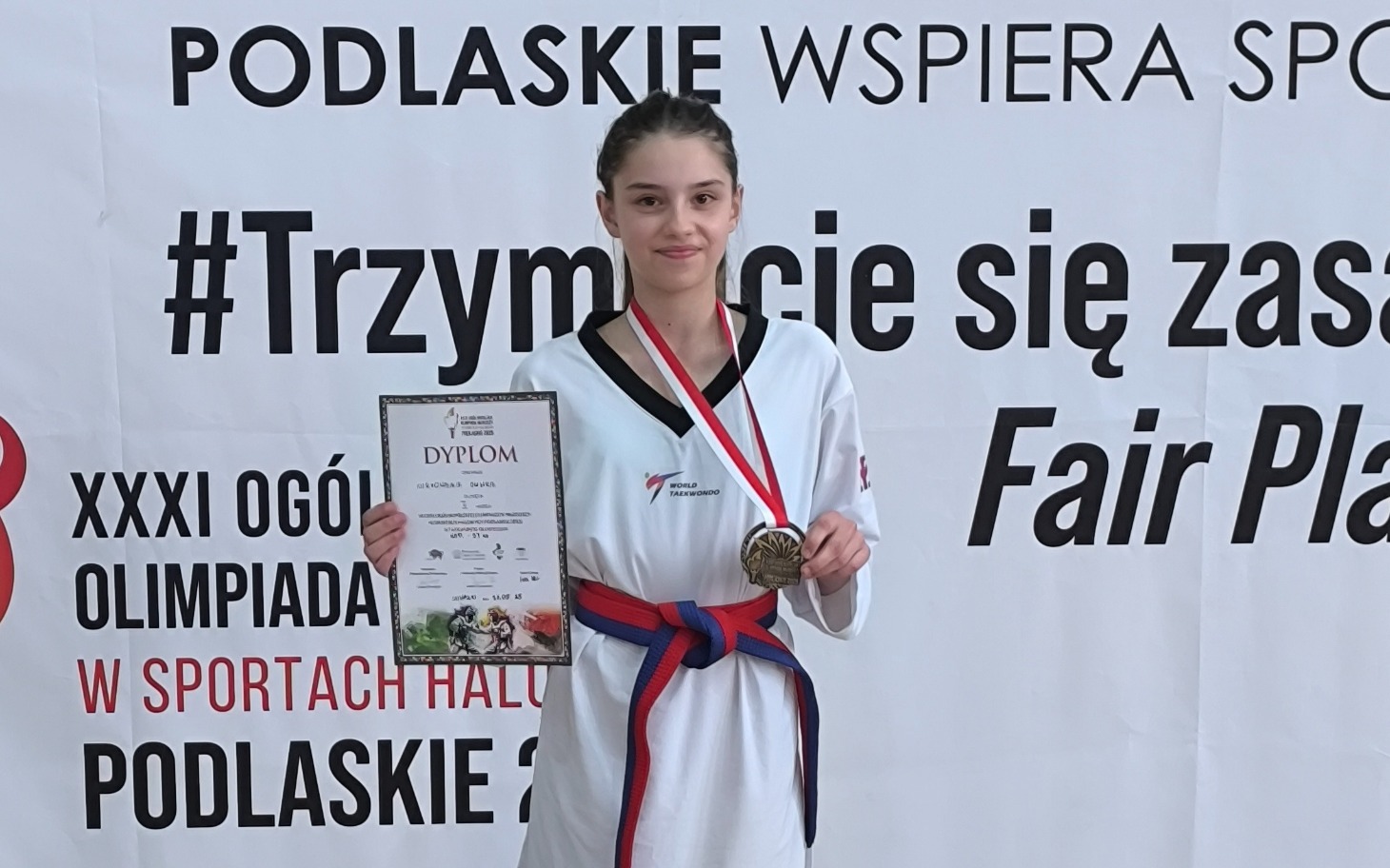 Młoda zawodniczka z dyplomem i medalem, dumnie prezentująca osiągnięcia.