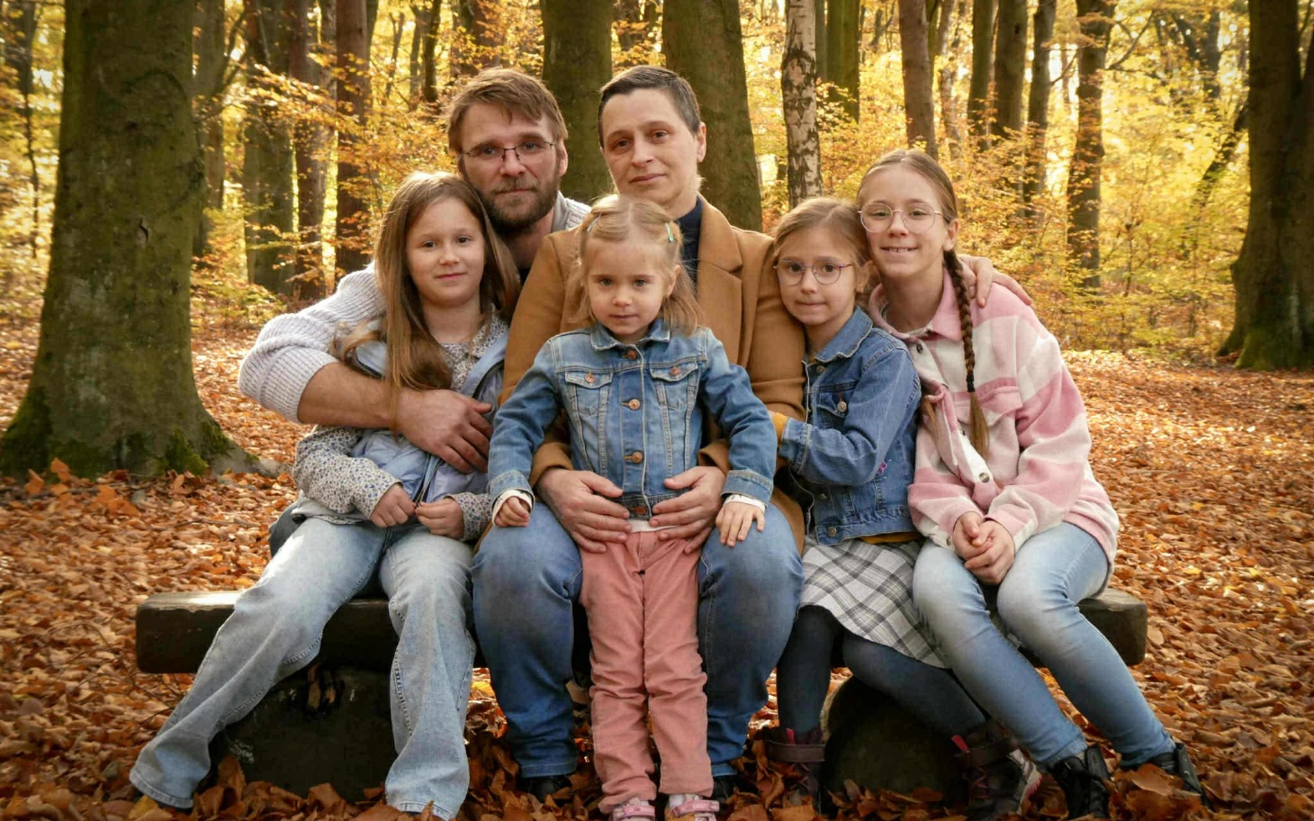 
        Rodzina na ławce w lesie, otoczeni jesiennymi liśćmi.
    