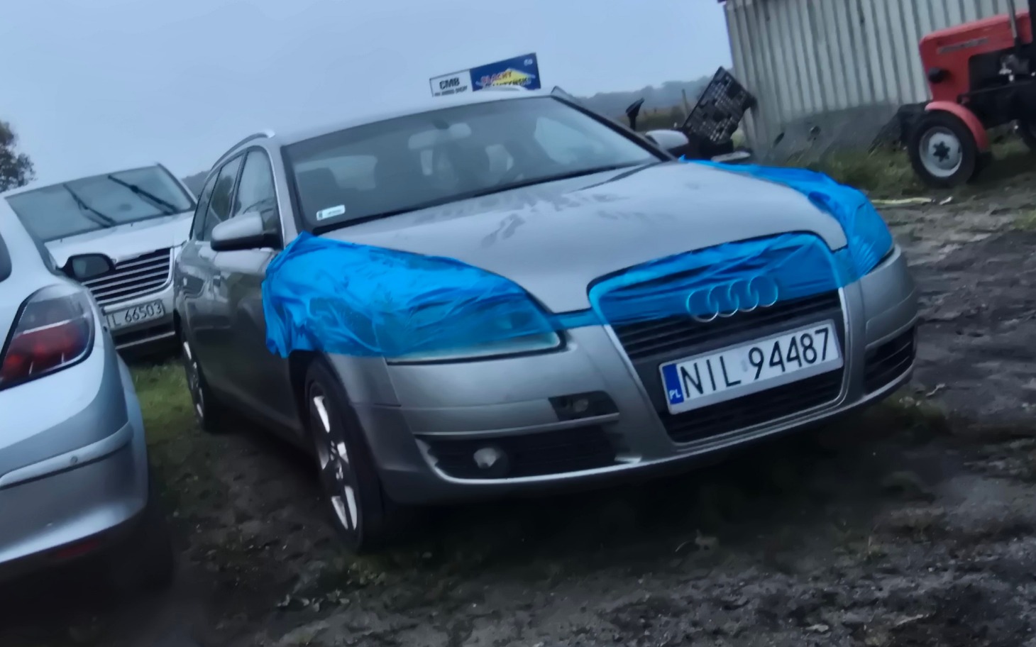 
        Samochód z niebieską folią na masce, zaparkowany na podwórku.
    