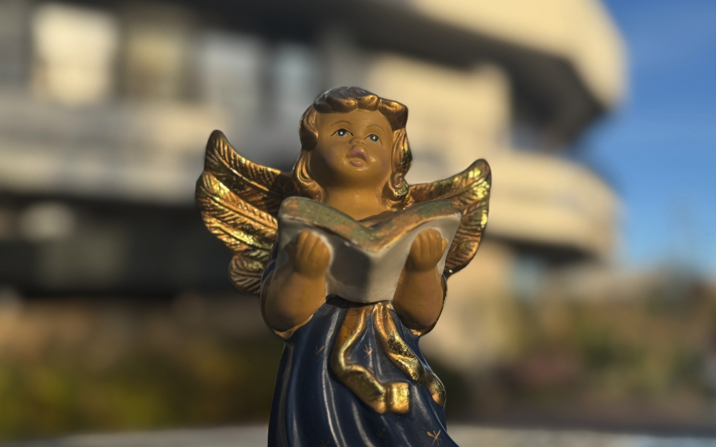 
        Figurka aniołka trzymającego książkę na tle budynku.
    