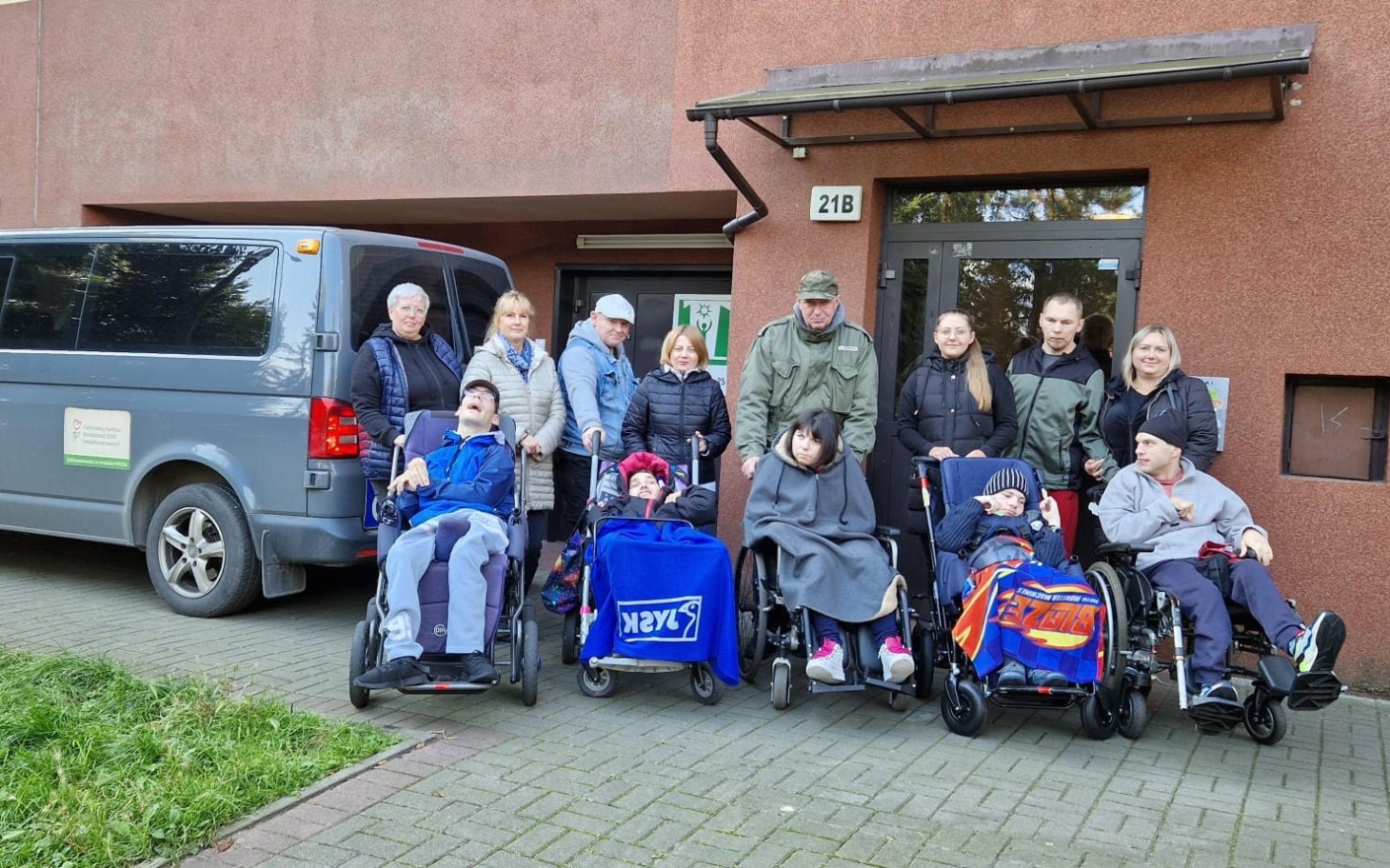 
        Grupa osób z niepełnosprawnościami przed budynkiem, wózki inwalidzkie.
    