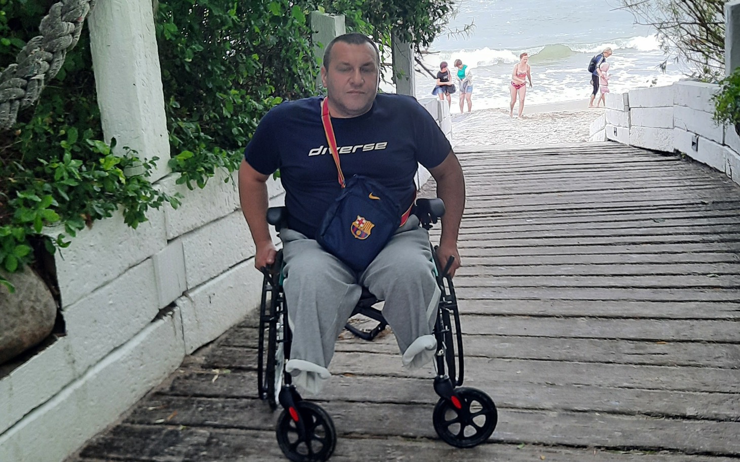 
        Mężczyzna na wózku inwalidzkim na plaży, w tle ludzie.
    