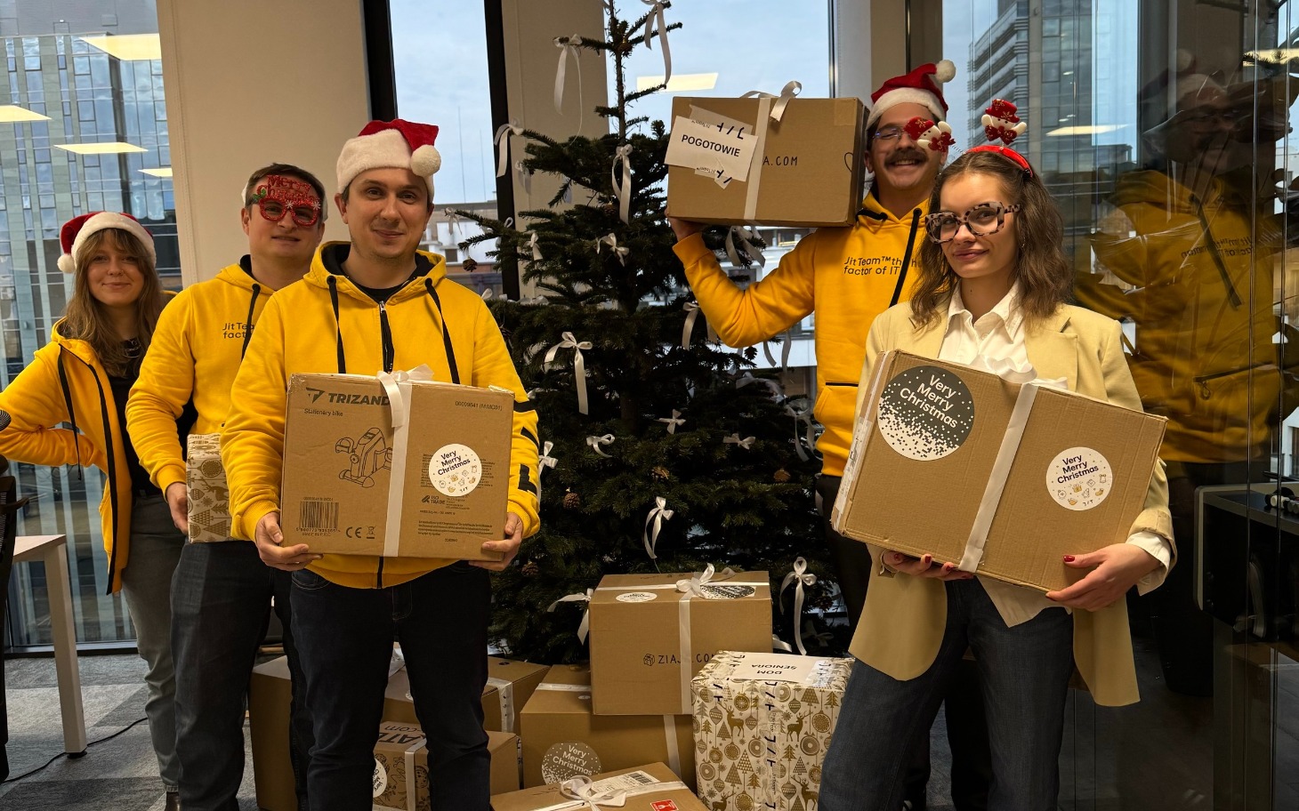 
        Grupa ludzi w żółtych bluzach z prezentami przy choince.
    