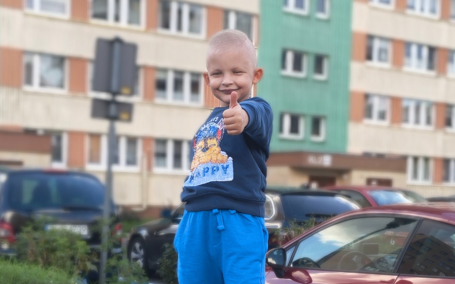 
        Chłopiec z uśmiechem i kciukiem w górze, stojący na parkingu.
    