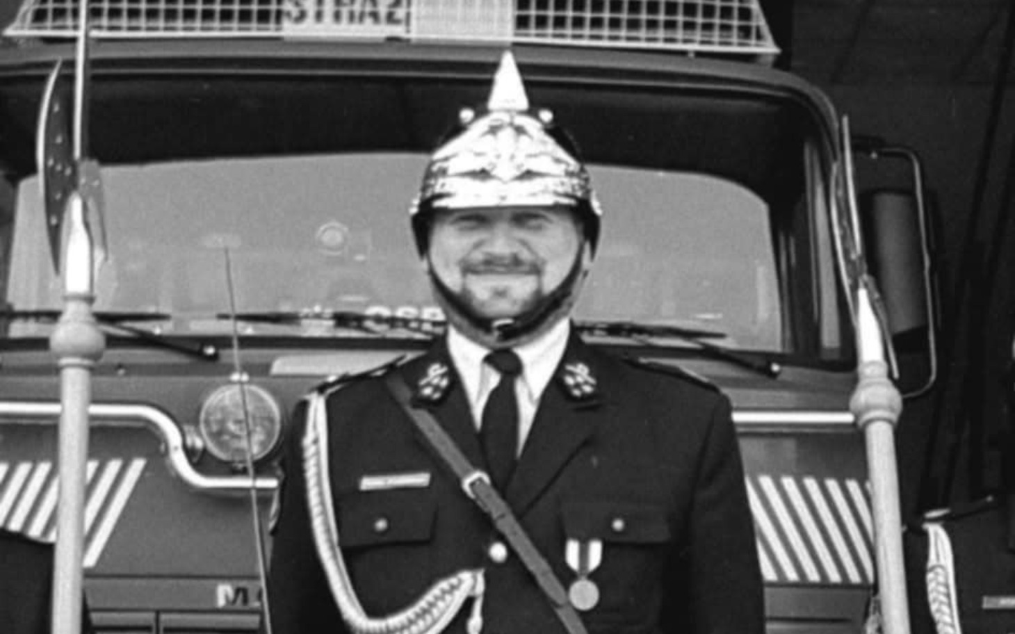 
        Strażak w mundurze, uśmiechający się przed wozem strażackim.
    