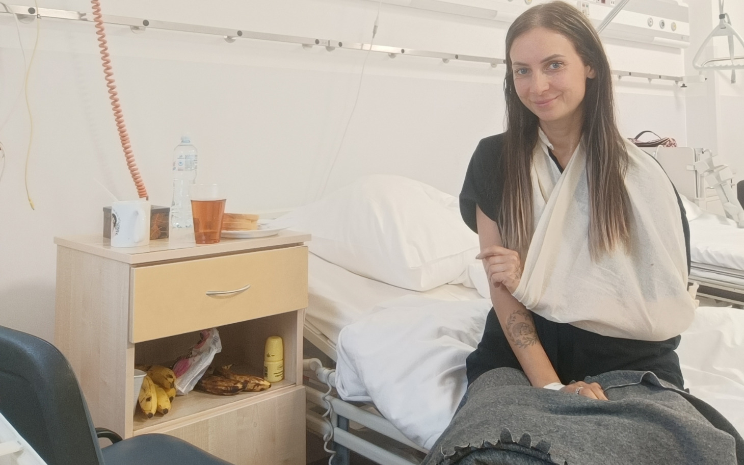 
        Kobieta w szpitalu, uśmiechająca się z unieruchomioną ręką.
    