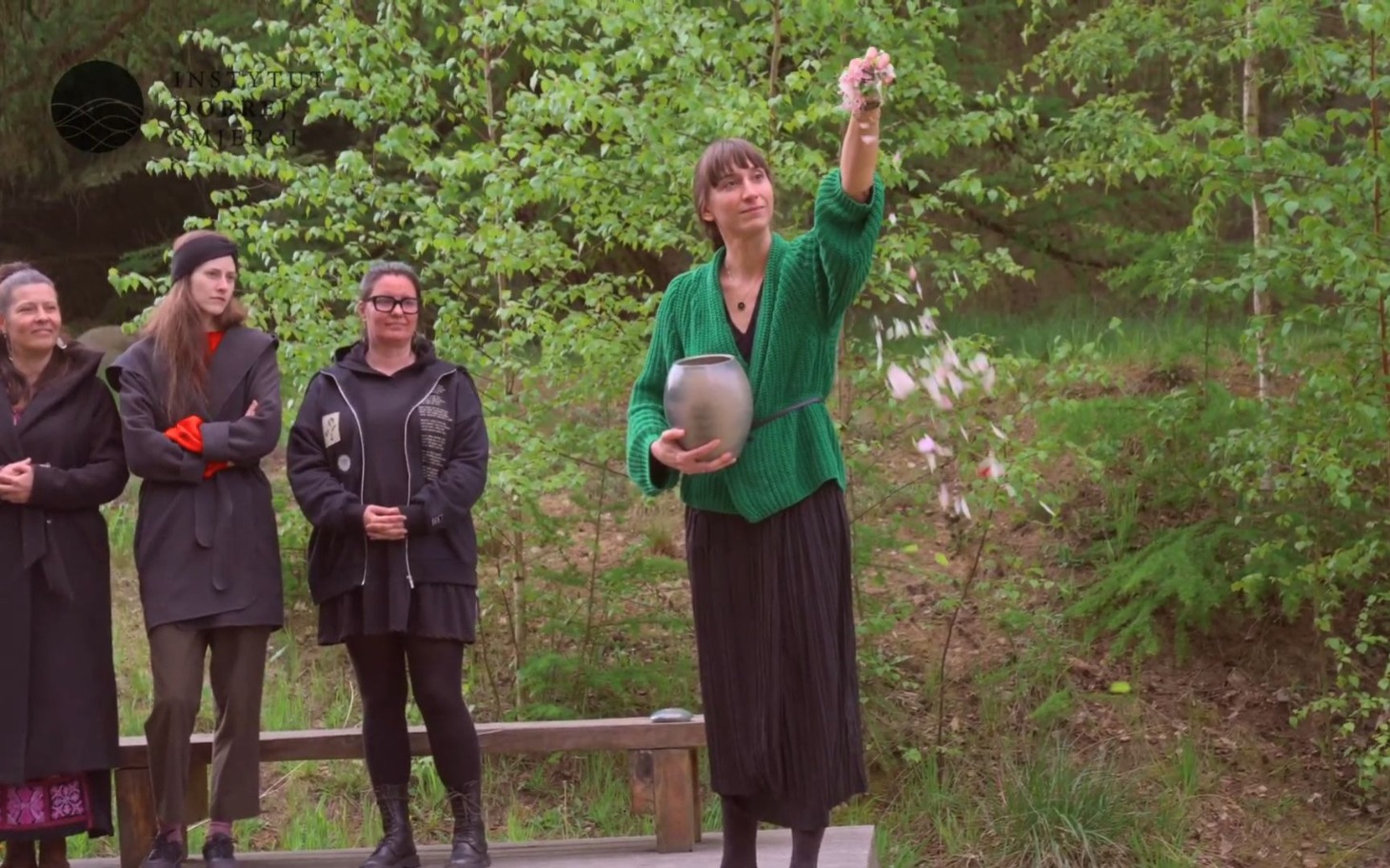 
        Kobieta w zielonym swetrze trzymająca naczynie, otoczona drzewami.
    