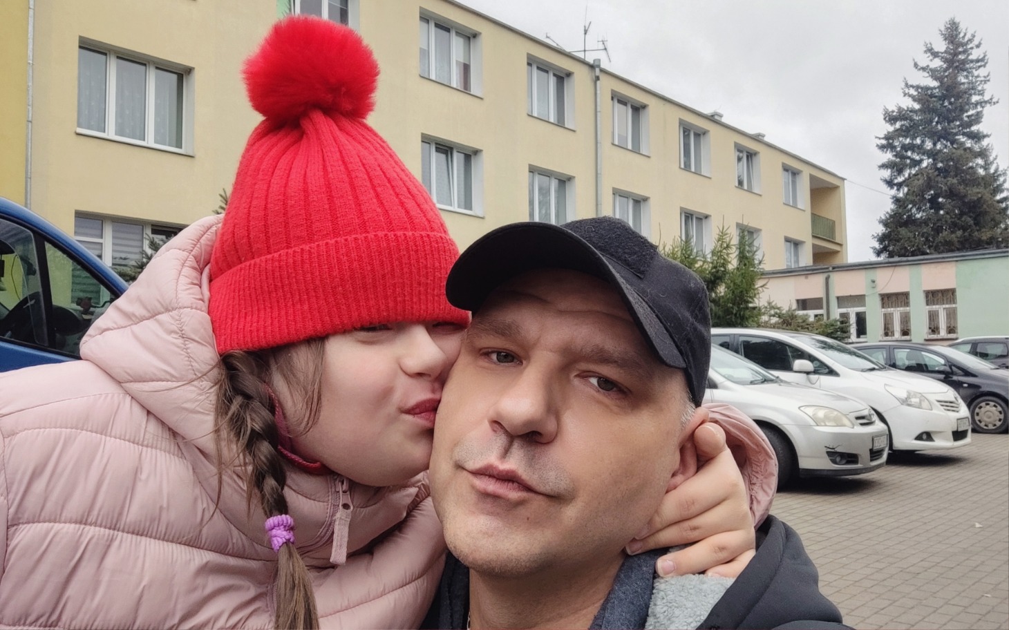 
        Dziecko w czerwonej czapce całuje dorosłego na tle budynków.
    